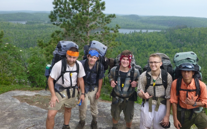 Five people wearing backpacks smile for a photo. They are standing on a rocky lookout, high above a green wooded area. In the distance, there is a body of water. 